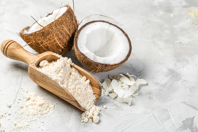 healthy coconut flour in wooden spoon next to opened coconut on table 